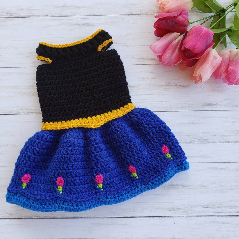 Puede incluir: Un vestido de ganchillo para un perro o gato peque&ntilde;o. El vestido es negro con un ribete amarillo y una falda azul con flores rosas.