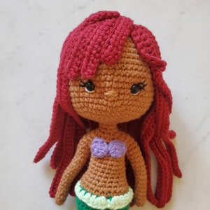 May include: Crocheted doll with red hair, brown skin, and a purple and green mermaid tail. The doll has black eyes and a crocheted top.