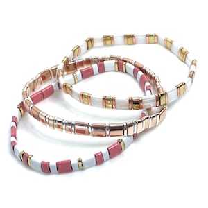 Puede incluir: Tres pulseras apiladas con cuentas blancas, rosadas y doradas. Las pulseras están hechas de cuentas pequeñas y cuadradas.