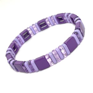 May include: A purple and lavender beaded bracelet with square and rectangular beads. The bracelet is made of plastic and has a simple design.