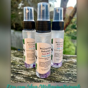 K&ouml;nnte beinhalten: Drei durchsichtige Glasflaschen mit schwarzen Spr&uuml;hk&ouml;pfen und Etiketten mit der Aufschrift "Wellspring Naturals Opal Mist Body Spray". Die Flaschen sind mit einer klaren Fl&uuml;ssigkeit gef&uuml;llt und haben am Boden violette Kristalle. Die Flaschen stehen auf einer Holzoberfl&auml;che.