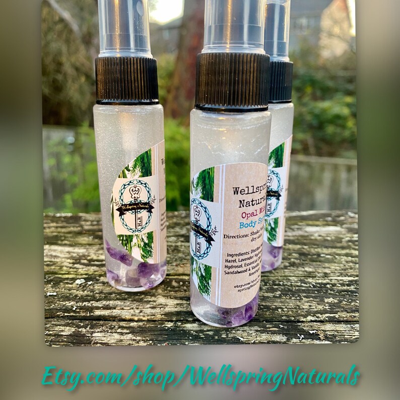 K&ouml;nnte beinhalten: Drei kleine, durchsichtige Glasflaschen mit schwarzen Spr&uuml;hk&ouml;pfen, gef&uuml;llt mit einer klaren Fl&uuml;ssigkeit und Amethyst-Kristallen. Die Flaschen tragen Etiketten mit der Aufschrift "Wellspring Naturals Opal Mist Body Spray" und Gebrauchsanweisungen.
