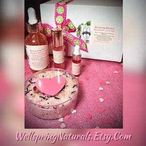 May include: A gift set with a pink heart-shaped bath bomb, a bottle of body oil, a bottle of essential oil, and a bottle of perfume. The set is packaged in a white box with a pink and green ribbon. The text on the box reads "Wellspring Naturals Abundant Love Set".