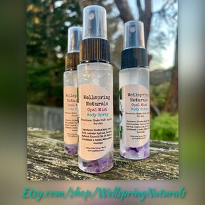 K&ouml;nnte beinhalten: Drei durchsichtige Glasflaschen mit schwarzen Spr&uuml;hk&ouml;pfen und Etiketten mit der Aufschrift "Wellspring Naturals Opal Mist Body Spray". Die Flaschen sind mit einer klaren Fl&uuml;ssigkeit gef&uuml;llt und enthalten am Boden kleine lila Kristalle. Die Etiketten enthalten auch Gebrauchsanweisungen und eine Zutatenliste.