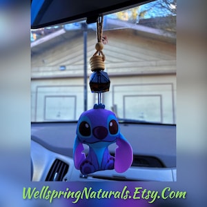 May include: Blue and purple Stitch character hanging from a rearview mirror with a small glass bottle filled with essential oil. The bottle is attached to a wooden bead and twine.