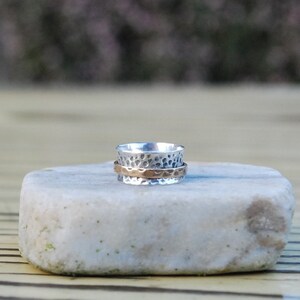 May include: A silver and copper spinning ring with a hammered texture. The ring is sitting on a light grey stone.