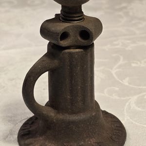 May include: A dark gray, weathered cast iron screw jack featuring a circular base with embossed text, a vertical cylindrical body with a side handle, and an adjustable threaded top component with a cross-shaped head for manual operation.