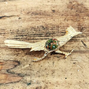 May include: A gold-toned metal brooch shaped like a roadrunner bird in a running pose. The bird features a textured feather design and a central oval-shaped cabochon stone with mottled green and reddish-brown patterns set into its body.