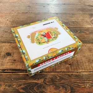 Könnte beinhalten: Eine geschlossene Romeo y Julieta Zigarrenkiste. Die rechteckige Kiste hat eine weiße Oberseite mit einer farbenfrohen Illustration und goldenen Akzenten. Die Kiste trägt die Aufschrift "Romeo y Julieta" und "Exhibition No. 3".