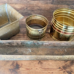 以下が含まれることがあります： さまざまな形のヴィンテージ真鍮プランターの詰め合わせ。正方形のプランター、ハンドル付きの小さなバケツ型プランター、赤いバンドが付いた円筒形のプランター、装飾的なエッジが付いた長い長方形のプランターが含まれます。すべて木製の表面に置かれています。