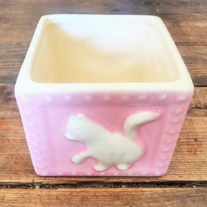 Puede incluir: Una maceta de cerámica rosa con una silueta de gato blanco en la parte delantera. La maceta tiene un interior blanco y un patrón de puntos alrededor del borde superior.