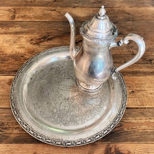 May include: A silver coffee pot with a decorative lid and handle sits on a matching round tray. The tray has an ornate border and engraved details. The set is displayed on a wooden surface.
