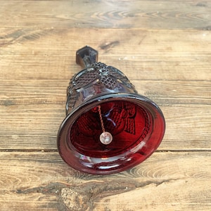 May include: A ruby red glass bell with a detailed floral pattern and a clear glass clapper. The bell is resting on a wooden surface. The bell's design includes a decorative handle and a detailed rim.