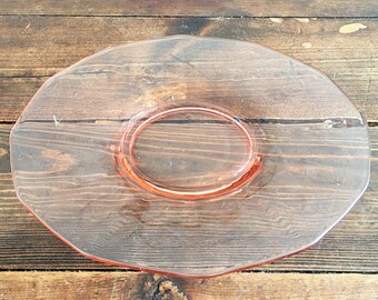 May include: A pink glass saucer with a scalloped edge. The saucer is empty and is sitting on a wooden surface.