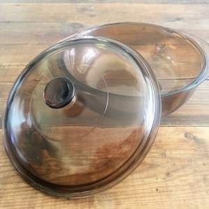 Könnte beinhalten: Eine klare, braune Auflaufform aus Glas mit passendem Deckel. Der Deckel hat einen dunkelbraunen Knopf. Die Form und der Deckel stehen auf einer Holzoberfläche. Die Form ist zum Kochen und Servieren bestimmt.