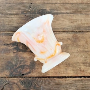 Op de afbeelding: Een vintage, wit en perzik kleurige glazen vaas met een uitlopende bovenkant en een voetstuk. De vaas heeft een delicaat, gedraaid patroon met oranje accenten. Twee kleine decoratieve handvatten zitten aan de zijkanten. De vaas staat op een houten oppervlak.