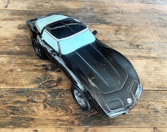 May include: A black Corvette model car with a white stripe on the roof. The car is sitting on a wooden surface.