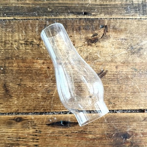 May include: A clear glass lamp chimney with a bulbous midsection that tapers toward a narrow top opening and a wider base opening. The transparent glass is smooth and features a simple, functional design intended for use with oil lamps.