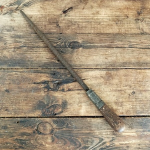 May include: An antique metal rasp with a wooden handle. The rasp is long and tapered, with a textured surface for filing. The handle is a light brown color, and the metal has a worn, aged appearance. The tool rests on a weathered wooden surface.