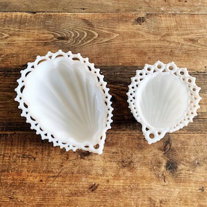 May include: Two white milk glass dishes with a teardrop shape and a scalloped, openwork lace-like border. The interior of each dish features a ribbed, shell-like pattern radiating from the base toward the decorative, intricate edges.