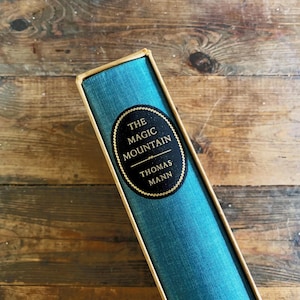 Puede incluir: Un libro de tapa dura titulado "The Magic Mountain" de Thomas Mann. El libro tiene una cubierta turquesa con una etiqueta ovalada negra y dorada. El libro está en una caja beige, sobre un fondo de madera.