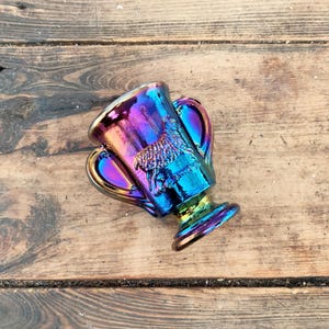May include: Iridescent glass trophy cup with two handles. The cup features a relief design of a ram. The colors shift from purple to blue and green. The trophy is set on a wooden surface.