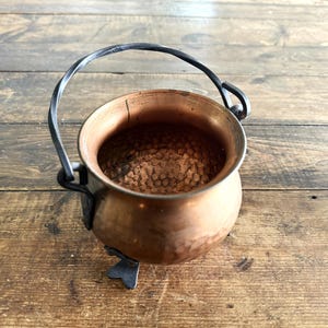 May include: A small, round copper cauldron featuring a hammered texture on the exterior and interior. The vessel is supported by three dark metal feet and includes a matching dark metal arched handle attached to the sides with looped brackets.