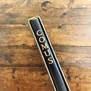 May include: A black book spine with the word COMUS printed in gold lettering. The book is encased in a light-colored, metallic-toned slipcase that frames the spine. The spine has a textured surface and shows signs of wear at the top edge.