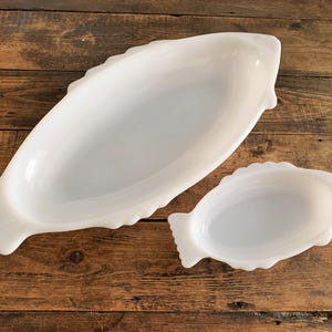 Può includere: Due piatti da portata bianchi a forma di pesce. Il piatto più grande è ovale e il più piccolo è una versione in miniatura. Entrambi hanno una finitura lucida e sono appoggiati su una superficie di legno rustico.