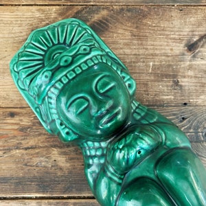 May include: A glossy, emerald green ceramic sculpture of a reclining figure with a detailed headdress. The figure has closed eyes and a serene expression. The sculpture is placed on a rustic, wooden surface.