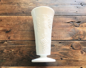May include: A white, vintage-style vase with a flared top and a pedestal base. The vase features a raised grape and vine design. The vase is made of opaque glass and is approximately 23 cm tall.