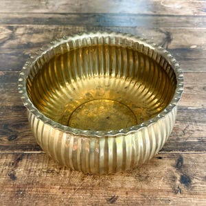 May include: A round, polished brass bowl featuring a vertical ribbed texture along its exterior and interior walls. The bowl has a scalloped, wavy rim edge that follows the pattern of the vertical fluting, creating a consistent metallic finish.