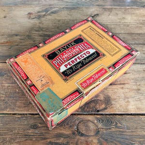 May include: Vintage Bayuk Phillies Perfecto cigar box. The box is rectangular with a tan and red color scheme. Text on the box includes "Bayuk Phillies Perfecto" and "It's Ripe Tobacco!" The box is on a wooden surface.