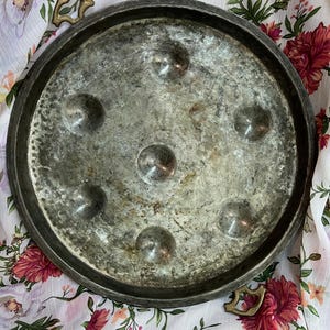 May include: A round, silver-colored metal tray with a hammered texture and six raised circles. The tray has two brass handles on the sides.