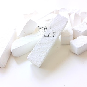 Könnte beinhalten: Eine Ansammlung weißer, rechteckiger, kreideartig strukturierter essbarer Tonstangen und -stücke. Auf der Oberfläche des mittleren Stücks befindet sich der aufgedruckte Text Snacks By Nature mit einer kleinen Blattillustration.