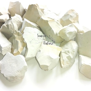 Peut inclure: Un tas de morceaux d'argile blanc cassé, de formes irrégulières. Les pièces varient en taille et en texture, certaines lisses, d'autres rugueuses. L'image contient le texte "Snacks By Nature".