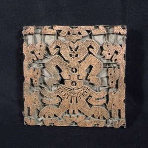 May include: A wooden printing block with a detailed, intricate design. The design features a central figure with a geometric pattern and is surrounded by smaller, repeating motifs. The block is made of a dark wood and has a worn, aged appearance.