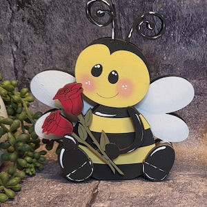 May include: A wooden bee figurine with a yellow body, black stripes, and white wings. The bee holds a stem with two red roses. The bee has a smiling face with rosy cheeks and decorative wire antennae.