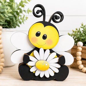 May include: A wooden bee figurine with a yellow head, black body, and white wings. The bee holds a white daisy with a yellow center. The bee has black antennae and rosy cheeks. The figurine is sitting on a wooden surface.