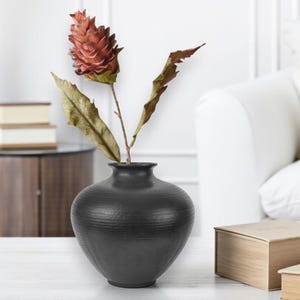 May include: A black, textured ceramic vase holds a dried flower arrangement. The arrangement features a reddish-brown flower and several dried leaves. The vase is round with a narrow neck and sits on a white surface. The background includes a white couch and wooden boxes.