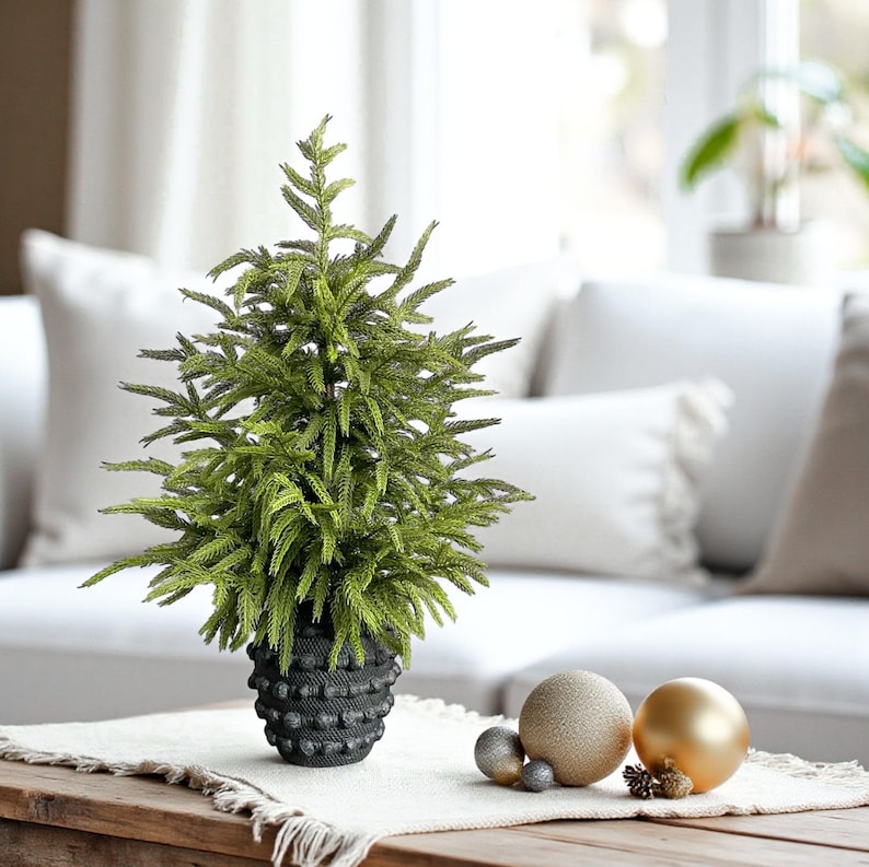 K&ouml;nnte beinhalten: Ein kleiner, k&uuml;nstlicher Weihnachtsbaum in einem strukturierten schwarzen Topf. Der Baum hat leuchtend gr&uuml;ne Nadeln und steht auf einem Holztisch mit dekorativen Ornamenten. Die Ornamente umfassen eine goldene, eine beige und zwei silberne Kugeln.