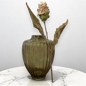 May include: A decorative olive green glass vase with vertical ribbed detailing. A single stem with a beige flower and two brown leaves is placed inside the vase. The vase is on a white marble surface.