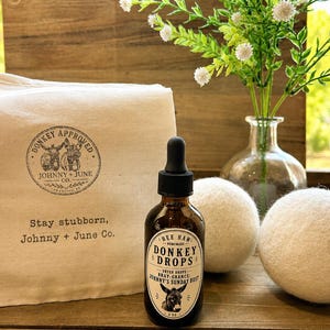 May include: A 2 oz amber glass dropper bottle labeled Donkey Drops with a donkey illustration, positioned next to a beige canvas drawstring bag featuring a Donkey Approved logo and the text Stay stubborn, Johnny + June Co., and two white wool dryer balls.