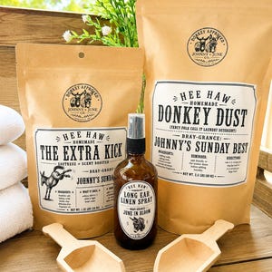 May include: Two brown paper pouches labeled Hee Haw Homemade Donkey Dust and The Extra Kick, an amber glass spray bottle labeled Long Ear Linen Spray, and two wooden scoops. The labels feature a donkey logo and the text Johnny + June Co.