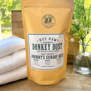 May include: A tan stand-up pouch of laundry detergent labeled Donkey Dust with a circular logo featuring two donkeys. The label includes the text Hee Haw Homemade Donkey Dust, Johnny's Sunday Best, and states a net weight of 2.5 lbs (40 oz).