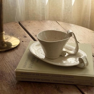 May include: A cream-colored ceramic cup and matching oval saucer with a speckled finish. The cup features a distinctively curved, scroll-like handle, and the saucer includes a matching scroll-like detail on one side, resting on two stacked books.