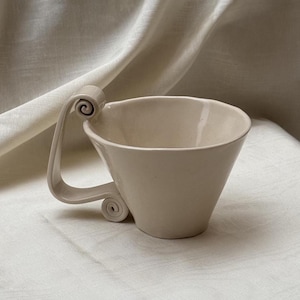 May include: A cream-colored ceramic cup with a wide, flared rim and a distinctive handle featuring two decorative scroll-like curls at the top and bottom attachment points. The cup has a smooth, glossy finish and an irregular, organic shape.