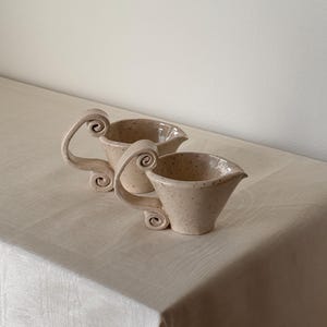 May include: Two speckled ceramic cream pitchers with decorative scroll handles. The pitchers are beige with dark brown speckles and a flared spout. The handles have a unique, curled design. The pitchers are on a light beige tablecloth.