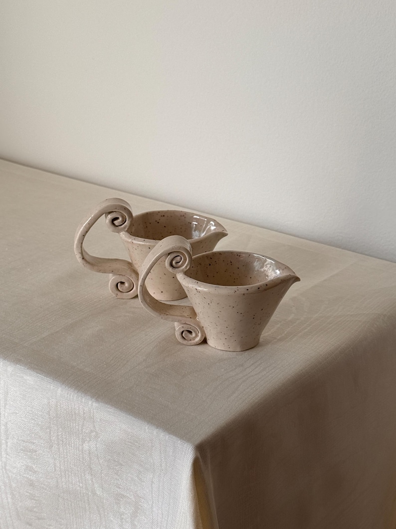 May include: Two speckled ceramic cream pitchers with decorative scroll handles. The pitchers are beige with dark brown speckles and a flared spout. The handles have a unique, curled design. The pitchers are on a light beige tablecloth.