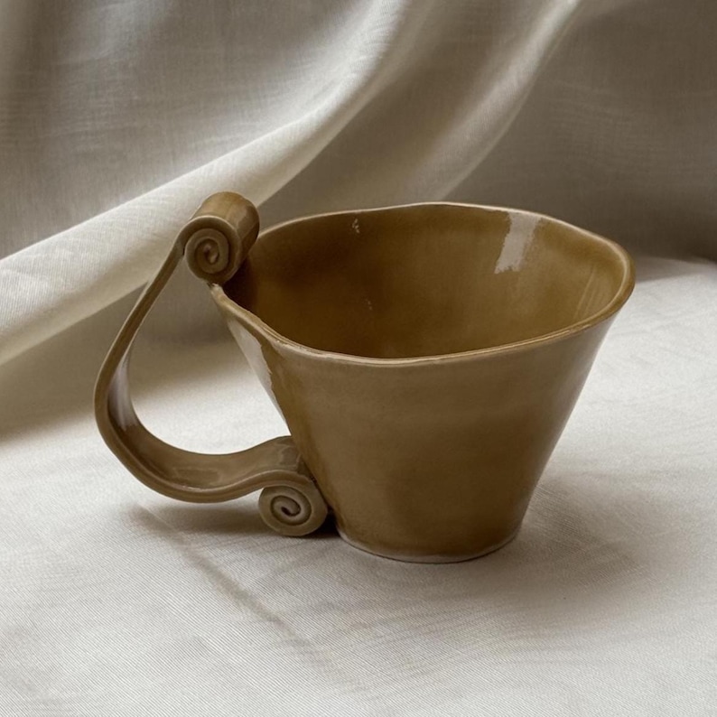 May include: A glossy, tan-colored ceramic mug with a wide, flared rim and a distinctive handle featuring two prominent, tightly coiled scrolls at the top and bottom attachment points, creating a unique silhouette with a smooth, reflective finish.